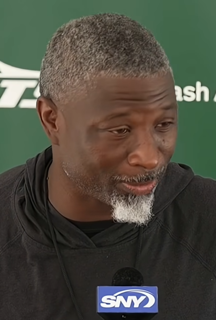 Jets head coach Aaron Glenn speaking at a news conference in front of a green Jets backdrop
