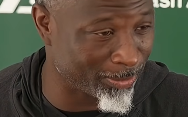 Jets head coach Aaron Glenn speaking at a news conference in front of a green Jets backdrop