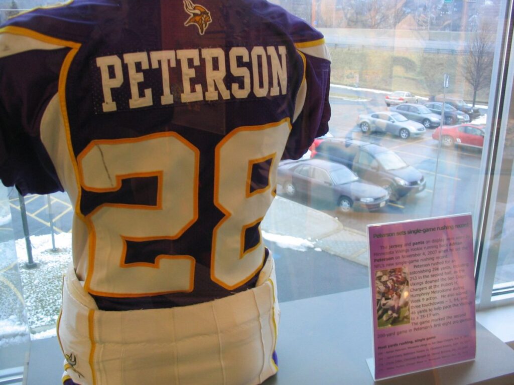 Adrian Peterson's purple Minnesota Vikings number 28 jersey and white pants on display in a glass case at the Pro Football Hall of Fame, next to a sign about his single-game rushing record