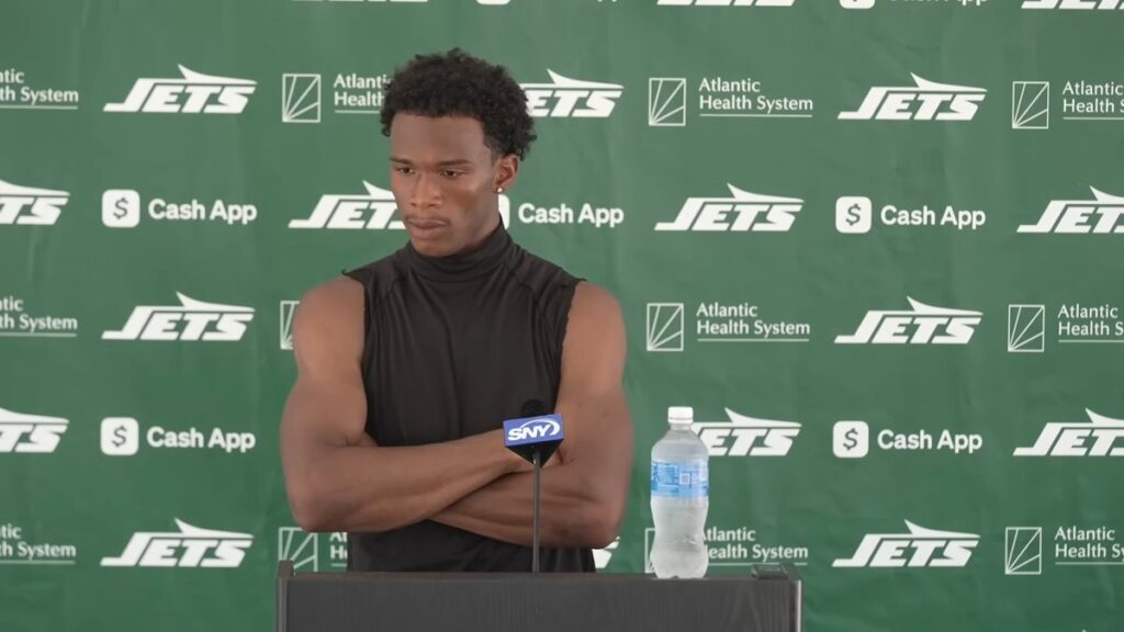 Garrett Wilson with his arms crossed at a podium in front of a green Jets backdrop