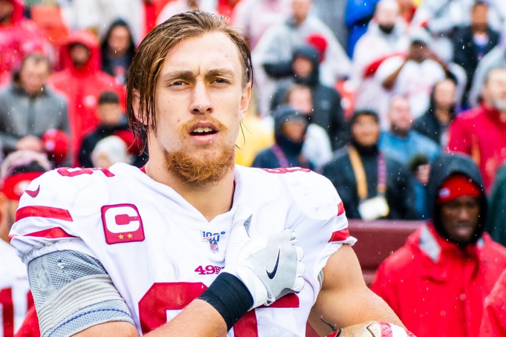 George Kittle in a white 49ers No. 85 jersey with a captain's patch, holding his helmet