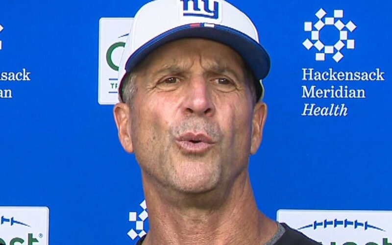 Giants head coach John Harbaugh in a white Giants cap speaking at a news conference in front of a blue sponsor backdrop