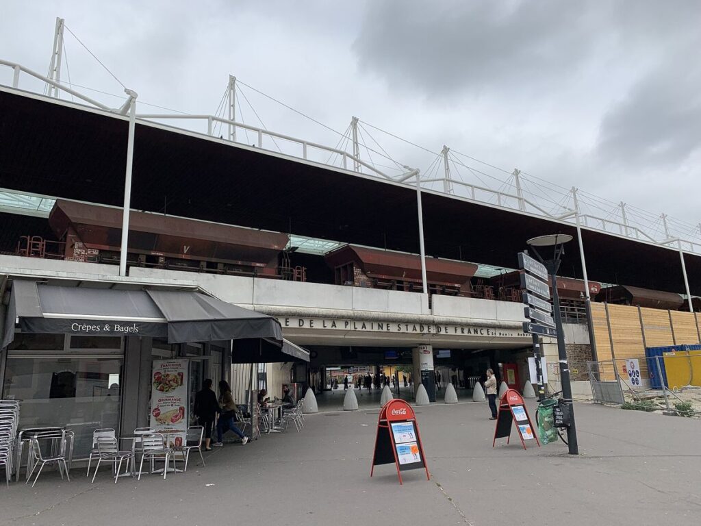 The entrance to the La Plaine Stade de France RER station in Saint-Denis under a grey sky, with a café terrace on the left