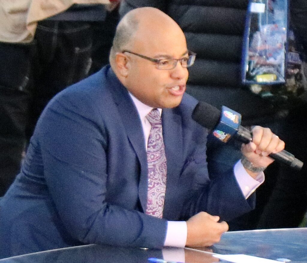 Mike Tirico speaking into an NBC microphone at a broadcast desk