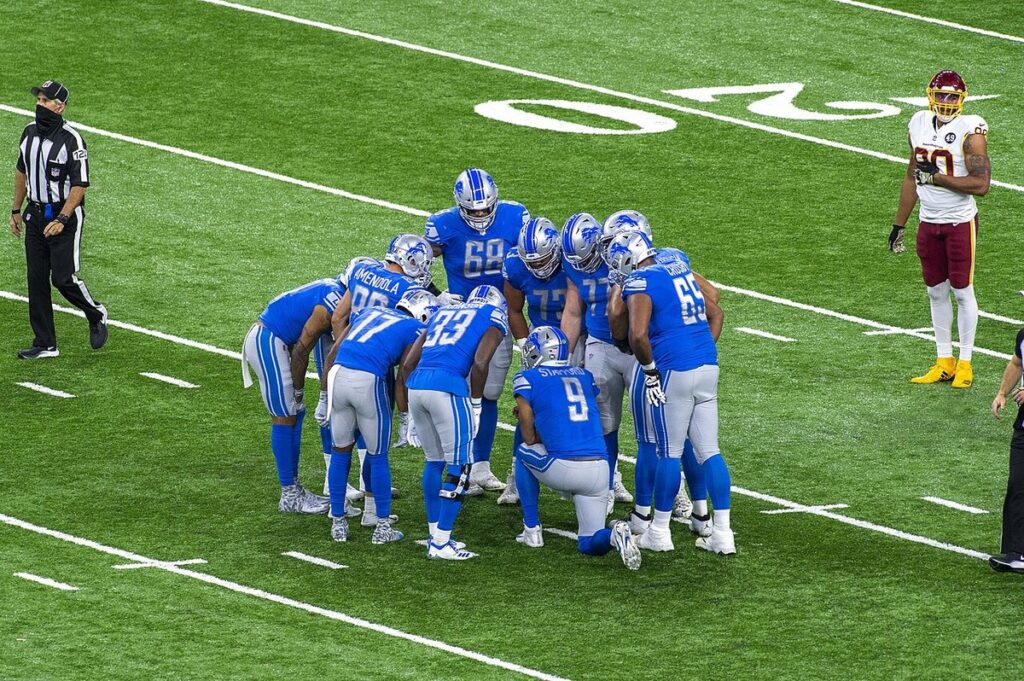 Detroit Lions offense kneeling in a huddle on the field at Ford Field while a game official and a Washington defender wait nearby