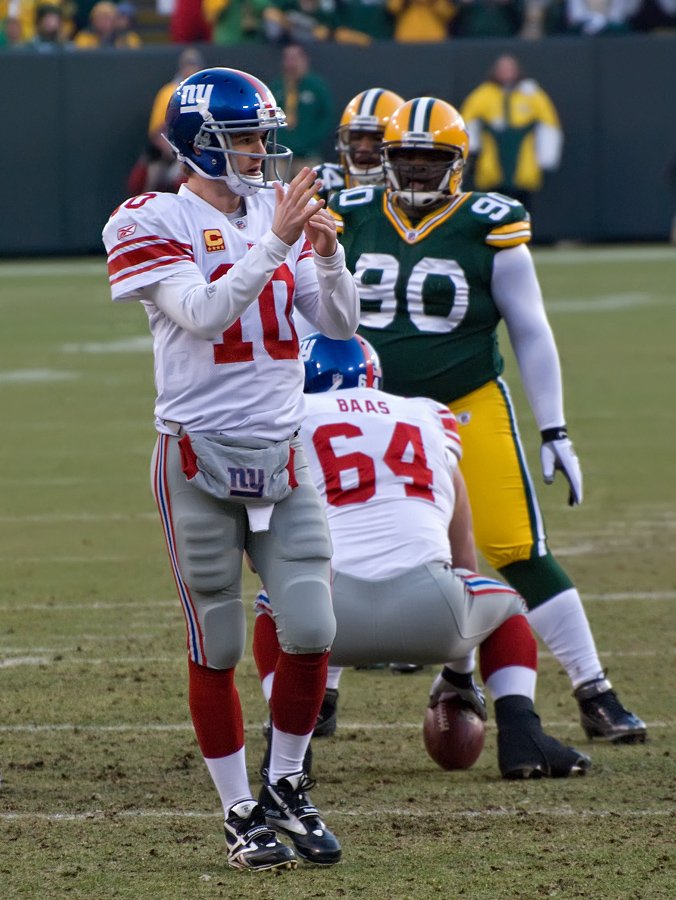 New York Giants quarterback Eli Manning in a white number 10 jersey making a T sign with his hands to call a timeout at Lambeau Field, with a Packers defensive lineman behind him