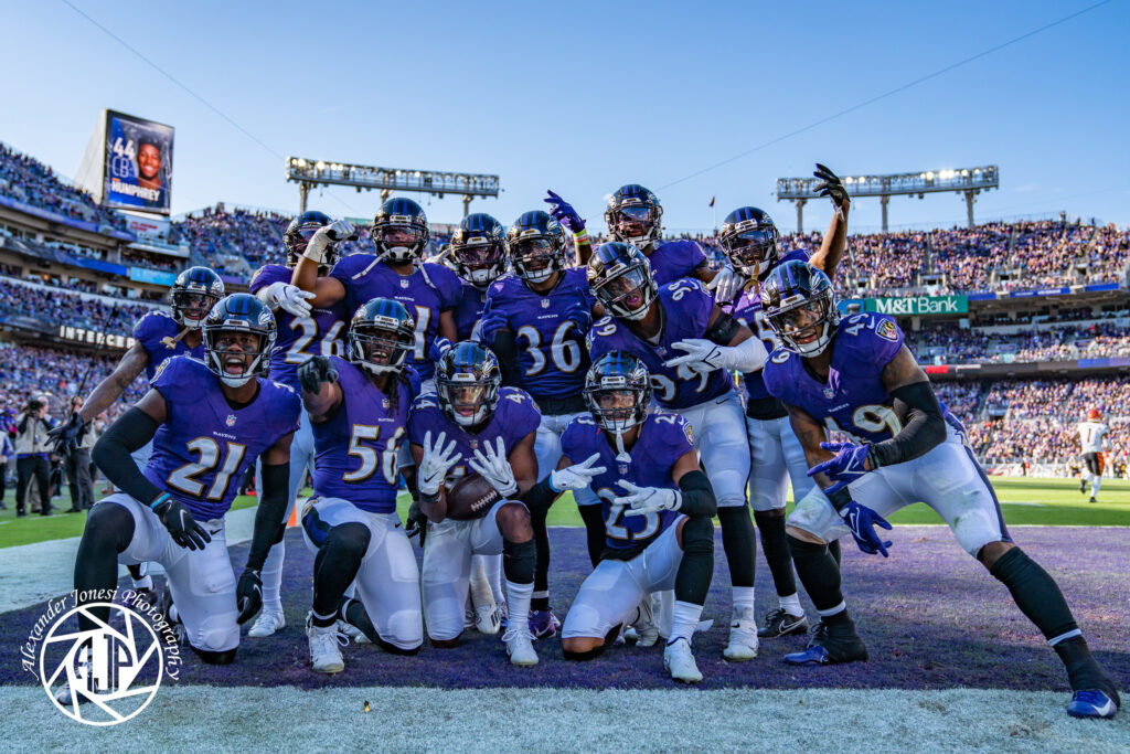 Marlon Humphrey, wearing No. 44, kneels with a football near the center of a group of Ravens players in purple uniforms.