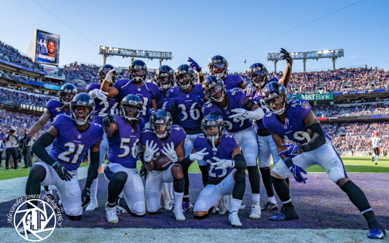 Marlon Humphrey, wearing No. 44, kneels with a football near the center of a group of Ravens players in purple uniforms.