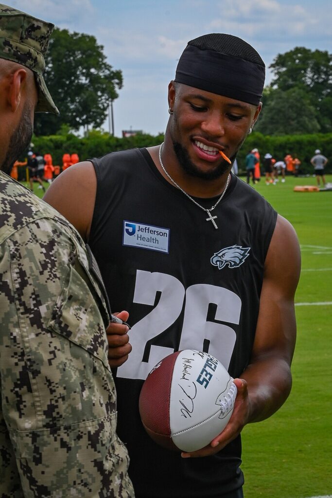 Saquon Barkley wearing an Eagles number 26 practice shirt and holding a football beside a service member at training camp in July 2025.