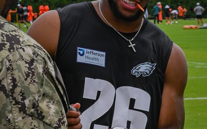 Saquon Barkley wearing an Eagles number 26 practice shirt and holding a football beside a service member at training camp in July 2025.